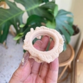 Elegant cream ruffled fabric scrunchie held against a natural green plant background - Nude Pink
