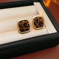 Detalle de pendientes de leopardo con borde dorado en un expositor acolchado blanco - Pendientes Cuadrados / café