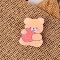 Cute orange acrylic bear brooch with pink heart pinned on brown woven fabric - Heart Hugging Bear
