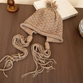Dettaglio del cappello in maglia color cammello con motivi a scacchi e lunghe nappe - Cappello in maglia con paraorecchie e nappe / Caffè Chiaro