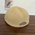 Plush beige beret for women featuring a metal tag on a textured wooden background - Camel