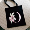 Black canvas tote bag with pink letter O and lily flower print on beige surface - Letter O / Black