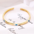 Detailed close-up of a gold-plated C-shaped bangle with blue enamel evil eye design - White