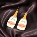 Close-up of champagne bottle earrings with pink labels and silver glitter on brown silk - Yellow Bottle (Silver Hook)