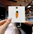 Dettaglio di una spilla a carota in smalto arancione con foglie verdi e bordi dorati - Carota