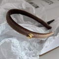 Nahaufnahme eines braunen Samtstirnbands mit goldenem M-Detail auf weißem Seidenpapier - Hellkaffee