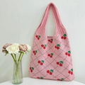 Pink knit tote bag with cherry and diamond patterns hanging on a white wall - Pink