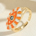Close-up of gold evil eye ring with orange enamel and blue crystal on beige surface - Orange