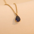 Golden drop necklace with deep blue gemstone pendant on a textured beige surface - October (Dark Blue)