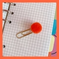Detalle de un clip dorado con pompón naranja sobre un cuaderno de cuadrícula - 8# Pompón Naranja