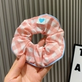 Pink checkered scrunchie with blue heart tag shown against a striped wall background - Large Grid / Pink