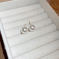 Close-up of elegant white pearl stud earrings with gold pins on a beige fabric surface - 12mm / Gold Plated White