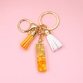 Detalle del llavero dorado con borla naranja y colgante de resina sobre fondo rosa - Letra I