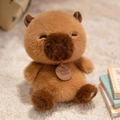 Cute sitting capybara plush toy in light brown with a wooden tag on a warm rug - Sitting Capybara