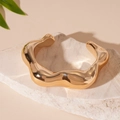 Detalle de brazalete dorado ondulado sobre piedra blanca con iluminación cálida y suave - Estilo Abierto Irregular / Dorado