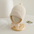 Macro detail of beige ribbed knit baby earflap hat with soft pom-pom and bow tie - 2-5 Years Old / Off White