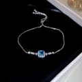 Detalle de una pulsera plateada con gema azul cuadrada y cristales sobre fondo negro - Pulsera-Plata-Azul
