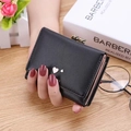 Women's black tri-fold mini wallet with gold heart clasp held by model over table - Black