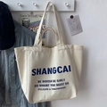 Detail of off-white canvas tote bag with blue bold text hanging in a lifestyle setting - SHAN (No Charm)