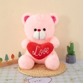Soft pink plush teddy bear sitting on a woven mat and holding a red heart with Love embroidery - Pink
