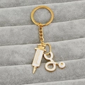 Gouden stethoscoop en spuit sleutelhanger met wit emaille op een grijs fluwelen blad - Goud