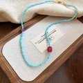 Dettaglio di collana boho con perline blu e gemma rossa su un vassoio di legno - Stile Perline Miste Blu e Rosa (HN9764)