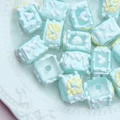 Macro view of light blue square hand-painted DIY beads with floral relief on a white tray - Trans Blue Square + Yellow Butterfly + White Cream