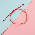 Detalle de pulsera de cordón trenzado rojo con barra rectangular dorada sobre fondo de color pastel - Cuerda Roja Oro Rosa