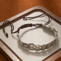 Close-up of ethnic braided bracelet with embossed silver metal charms on a wooden tray - 15 Flat Fish Alphabet Bracelet