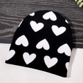 Berretto in maglia nera con motivi a cuore bianchi su sfondo di legno scuro - Taglia unica / Nero