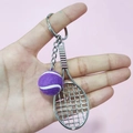 Close-up of a purple tennis ball and silver racket keychain held against pink background - 2cm Ball / Purple