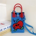 Detailed shot of a blue knitted apple bag with a red handle and magnetic closure on books - 5 Phone Bag-Red Apple Blue Base