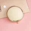 Round golden metal portable makeup mirror shown closed on a pink surface - 65mm / Rose Gold