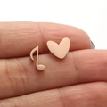 Rose gold asymmetrical heart and musical note stud earrings shown on fingers - Rose Gold