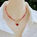 Collana artigianale di perline rosse e oro con ciondolo a cuore alato su camicia bianca - Collana di perline rosse con ciondolo a cuore alato