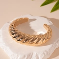 Detalle de un brazalete abierto dorado trenzado sobre un fondo de piedra con textura - Estilo Abierto Acanalado / Dorado
