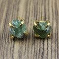 Close-up of raw green crystal stud earrings with gold four-claw setting on wood surface - Green Fluorite Style
