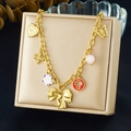 Dettaglio di una collana dorata con ciondoli cuore, fiore e fiocco su un cuscino beige - Stile Fiocco Colorato e Quadrifoglio (N3782)