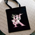Black canvas tote bag featuring a pink lily letter K floral print design - Letter K / Black