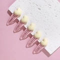 Close-up of five pink metal hair clips with yellow 3D hearts on a textured white block - Off White