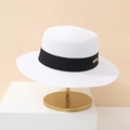 White woven flat top sun hat with black ribbon and gold metal plate on a stylish stand - White