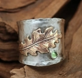 Detailed view of a wide silver band ring with a golden leaf and green gemstone on wood - US 6 / Oak Leaf Pattern with Green Stone (KYRA01487)