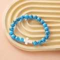 Blue turquoise style beads bracelet with a white cross charm on a beige textured tray - Blue Turquoise White Cross Bracelet