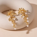 Detalle de pendientes de flor dorados con perlas blancas sobre soporte de cerámica beige - Pendientes de Botón de Flor y Hoja Dorada (Pin de Plata)