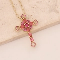Gold cross necklace with red and pink crystals on a textured beige stone slab - Red