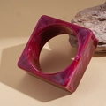 Dettaglio di un bracciale quadrato in resina marmorizzata rosa accanto a un pezzo di legno - Bracciale quadrato con motivo marmorizzato magenta
