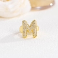 Close-up of golden balloon letter M ring with pavé cubic zirconia on soft floral background - M