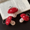 Three handmade red mushroom wool felt keychains with smiling faces on a book - Mushroom Trio