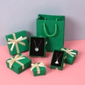 Macro view of dark green jewelry gift boxes with gold ribbons and matching carry bags - 7*7*3cm / Green