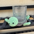 Macro shot of a handmade crochet turtle coaster holding a textured glass on a shelf - Knitted Coaster / Brown Green